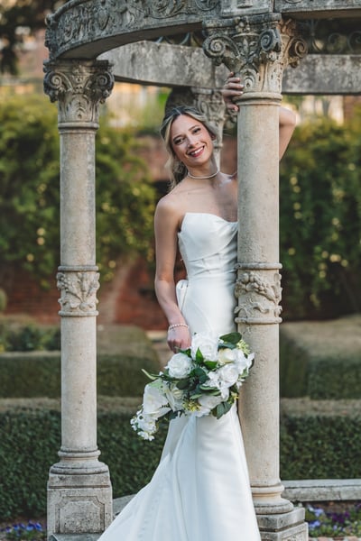 Bride smiling inside the ornate garden gazebo, bouquet in hand, afternoon light and boxwood hedges behind her