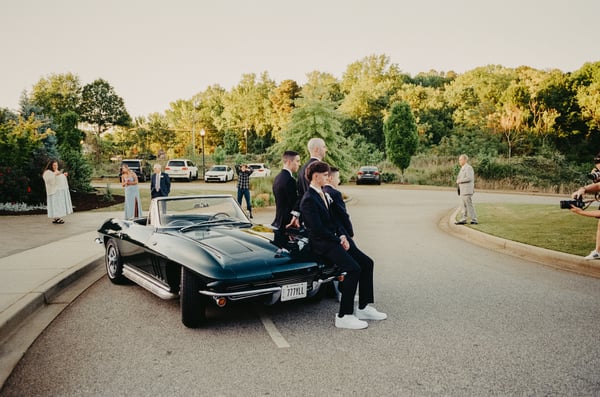 Junior groomsmen in dark suits perched on the front of the vintage Corvette while guests linger in the background at golden hour