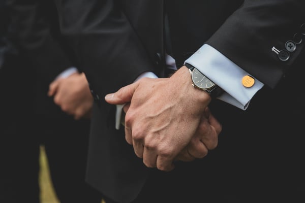 Close-up of the groom's clasped hands in a dark suit, wearing a watch and gold-monogrammed cufflinks, with a groomsman visible beside him