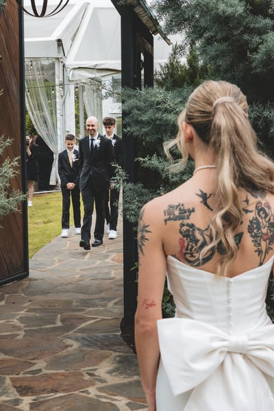 Wide view of the first look: bride in white gown facing groom in dark suit as he approaches through a garden doorway with young groomsmen watching
