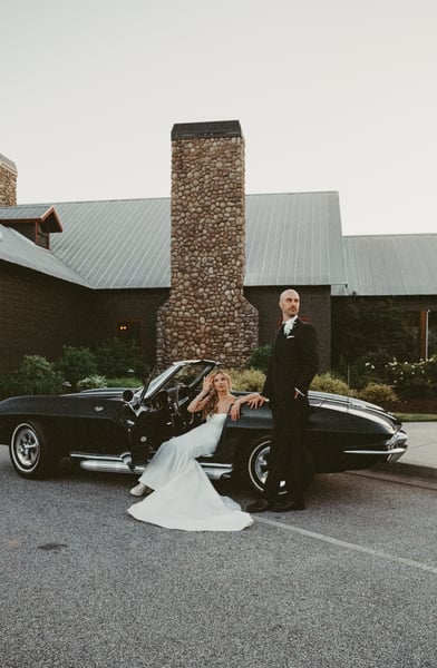 Bride leaning out of the open Corvette door with one hand raised to shield her eyes as the groom stands beside her, stone chimney venue behind