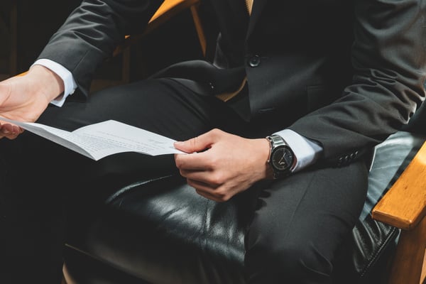 Groom in a dark suit reading his vows from a folded paper while seated, his watch visible on his wrist