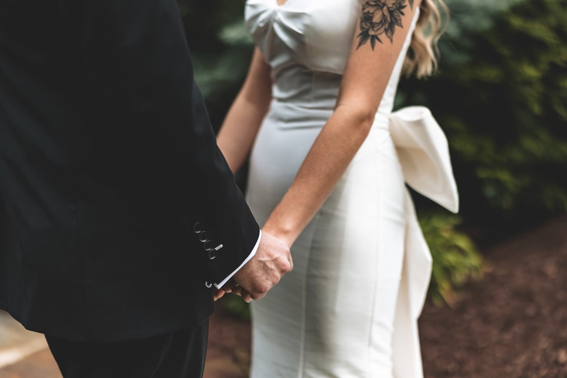 Close-up of the bride and groom holding hands, her arm tattoo and the gown's bow visible alongside his dark suit sleeve
