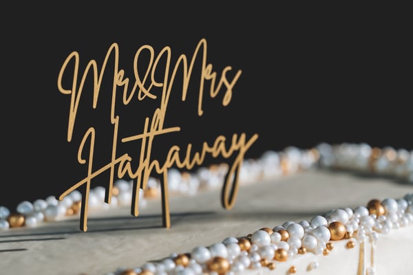 Close-up of the gold Mr. and Mrs. Hathaway cake topper against a dark background, with pearl and gold bead decorations along the cake edge