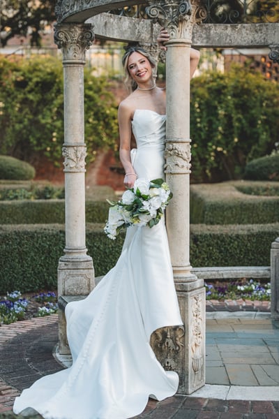 Bride in mermaid gown smiling inside the ornate garden gazebo, framed by stone columns, afternoon light and hedges behind