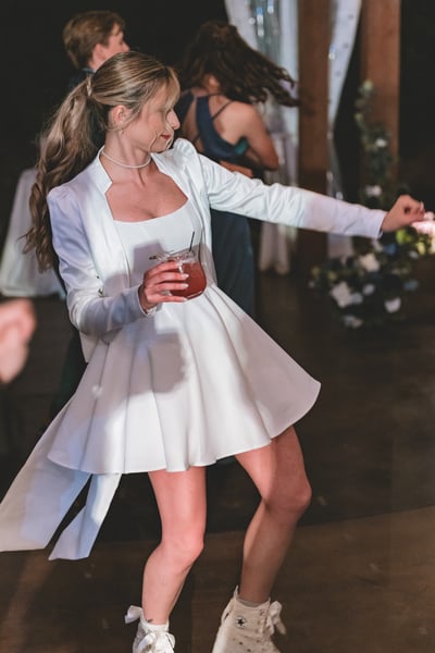 Overhead flat lay of end-of-night remnants — a half-eaten slice of wedding cake, a bottle of André Brut, the bride's pale blue bow clutch, white heart sunglasses, and a lace garter