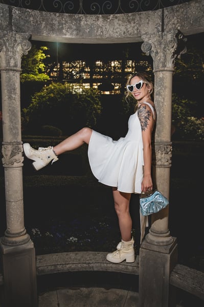 Bride in short white dress striking a playful pose inside the ornate iron-dome gazebo at night, one leg raised, blue bag, bejeweled Converse