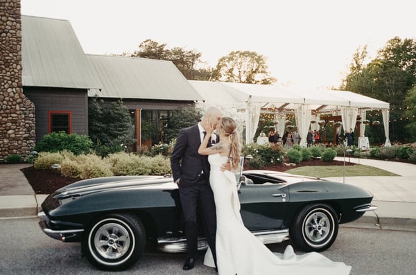 Bride and groom kissing leaned against the Corvette in front of the open reception tent, golden hour light across the venue
