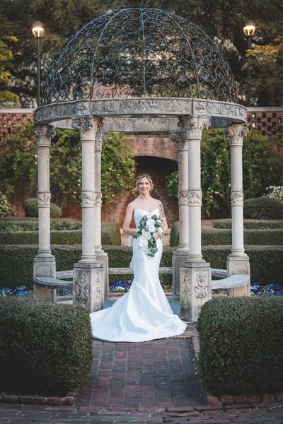 Bride standing centered in the iron-dome garden gazebo, facing camera, bouquet held at waist, smiling warmly with pansies and hedges behind