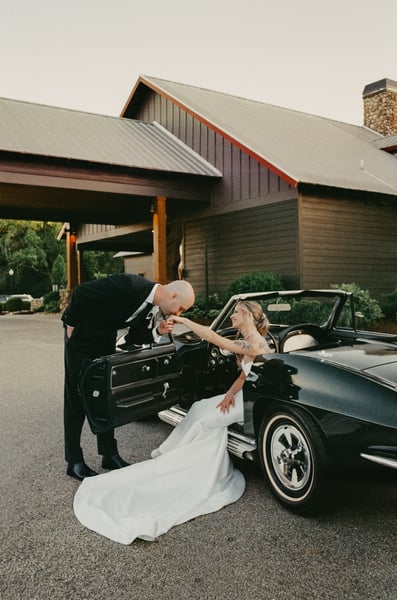 Groom bowing to kiss the bride's hand as she sits in the open Corvette door, her gown spread across the driveway