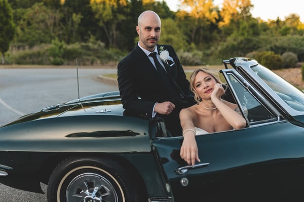 Front-on view of the classic Corvette with bride and groom kissing inside — his wedding band visible on the steering wheel in the foreground