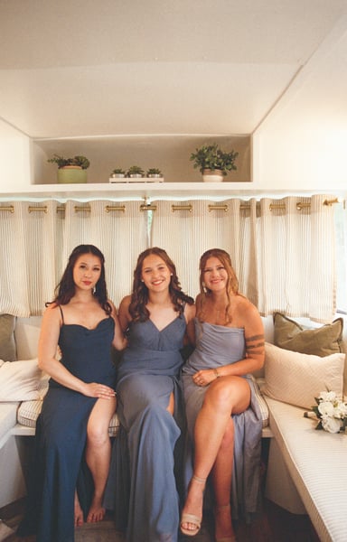 Three bridesmaids in navy and blue dresses seated together inside the airstream against a window of striped curtains