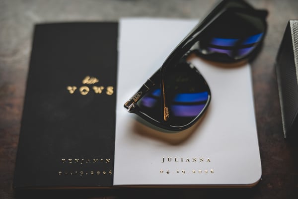 Close-up of two personalized vow books — black for Benjamin and white for Julianna — with a pair of monogrammed Persol sunglasses resting on top, all arranged on a wooden surface