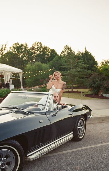 Bride standing in the Corvette in her wedding gown while groom sits at the wheel, string lights and tent in the distance