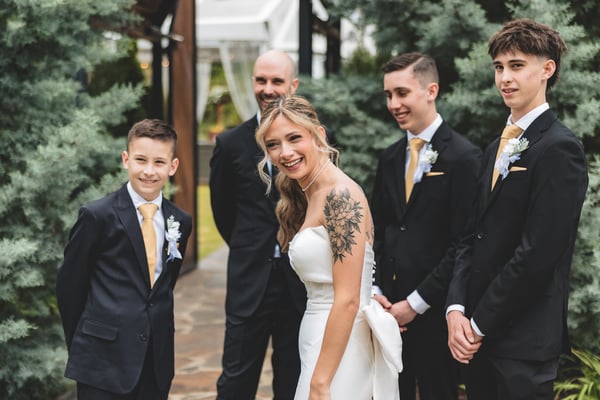 Bride in her white mermaid gown smiling brightly alongside the groom and three groomsmen in dark suits with gold ties, standing outdoors