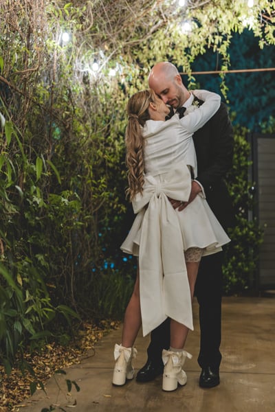 Bride and groom popping a champagne bottle together at night outdoors — champagne spraying dramatically as both laugh with delight
