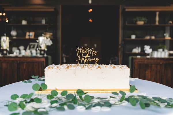 Rectangular white wedding cake topped with a gold laser-cut Mr. & Mrs. Hathaway cake topper and pearl-and-gold bead trim, displayed on a table with eucalyptus