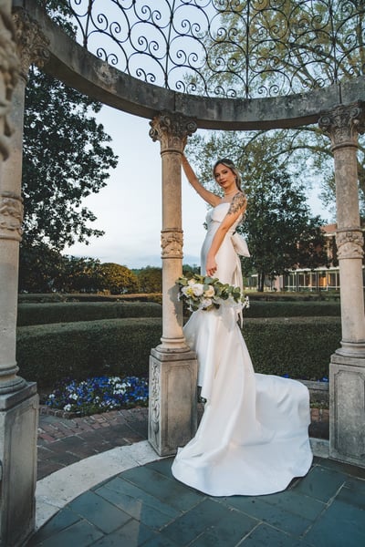 Bride standing inside a wrought-iron dome gazebo in the formal garden at twilight, bouquet at her side, reaching up to the ironwork