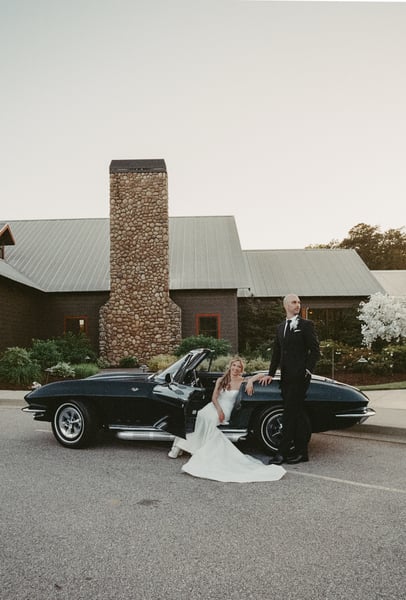 Bride seated in the open door of the Corvette with her gown spilling onto the pavement, groom standing tall beside her, chimney venue in the background