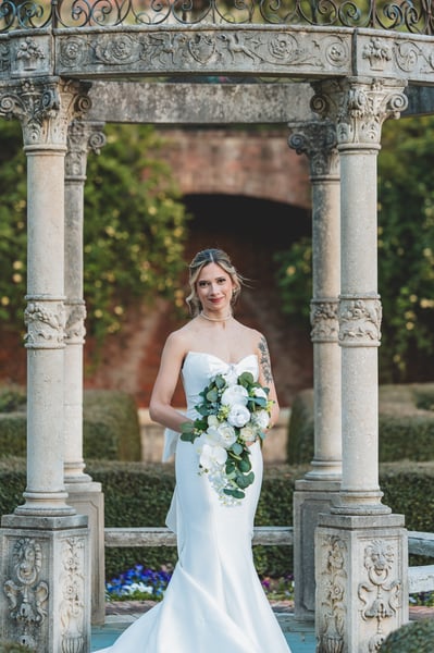 Bride standing in the ornate garden gazebo facing camera, bouquet held at waist, blue pansies and boxwood visible behind