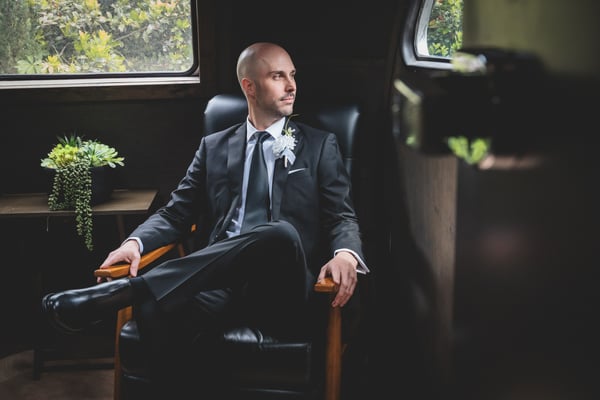 Groom seated in a leather chair inside a vintage aircraft cabin, gazing out the window with a white boutonniere on his lapel