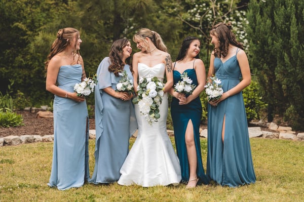 Bride in white strapless gown with bridesmaids in shades of dusty blue and teal, all laughing together on a garden lawn, each holding white and green bouquets