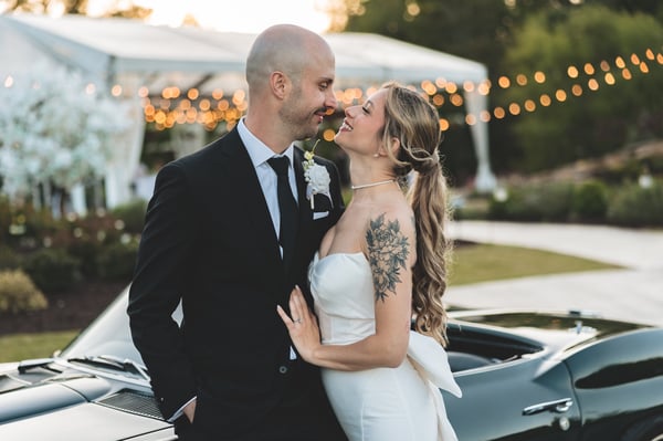 Bride and groom face to face in front of their vintage Corvette at dusk, string lights and white tent glowing behind them as they smile at each other