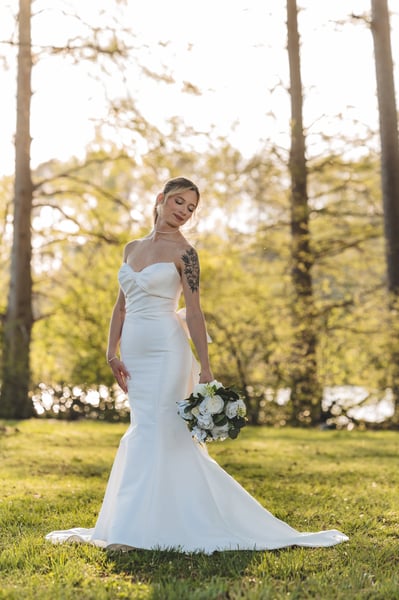 Full-length portrait of bride in open field, eyes gently closed, bouquet held at hip, surrounded by golden afternoon light and trees