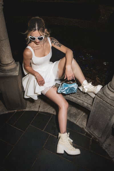 Bride in short white dress seated on stone bench at night in garden, looking down at bag and Converse, heart sunglasses perched on nose