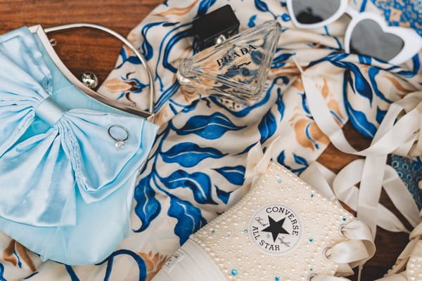 Flat lay of the bride's reception night accessories — a pale blue satin bow clutch, Prada perfume bottle, blue-and-white printed silk headscarf, bejeweled Converse sneaker, and white heart sunglasses