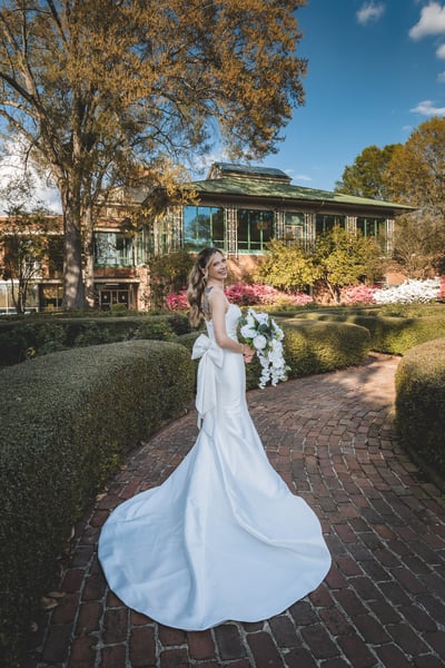 Bride turning to look back at camera in formal English garden with boxwood hedges, pink azaleas, and a glass conservatory in the background