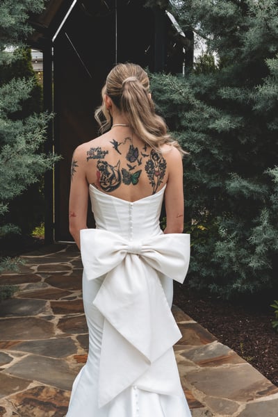 Back view of the bride in her white strapless mermaid gown with a large bow detail, revealing her back tattoos as she walks through a garden gate