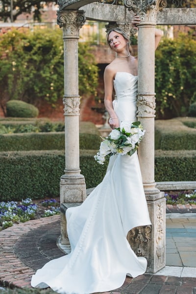 Bride standing between ornate stone columns of the garden gazebo, leaning back, bouquet held low, looking up