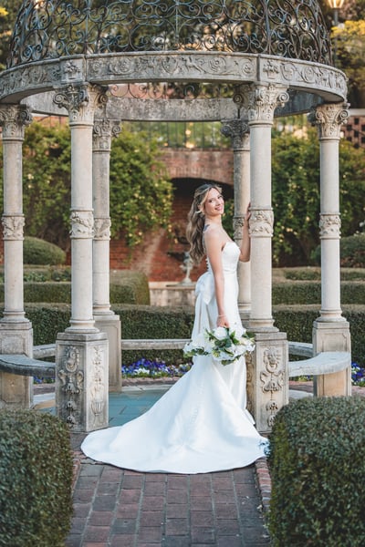 Bride partially turned away inside ornate iron-dome gazebo in the garden, gown train spread across the brick path