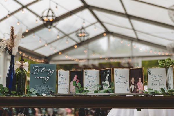 In loving memory display with framed photos of four loved ones who could not be there, arranged alongside greenery under string lights in a glass pavilion