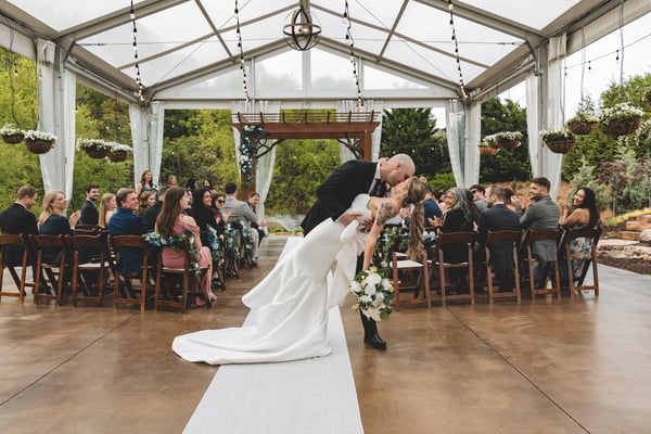 Groom dipping the bride for a dramatic first kiss at the altar beneath a glass-roofed pavilion, with guests seated on both sides and the wedding party looking on