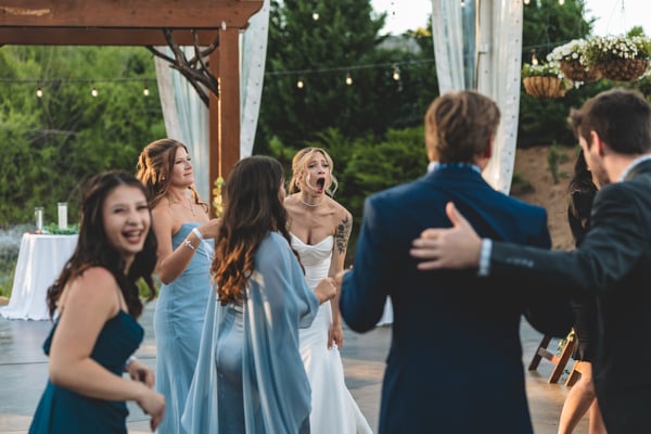 Bride in the middle of the dance floor surrounded by bridesmaids in blue and teal dresses, mouth open mid-song under the string lights