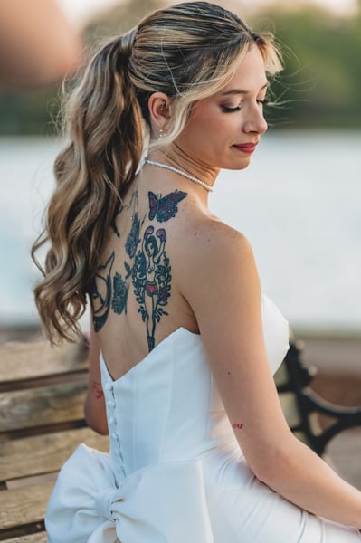 Bride seated on a bench at dusk, back to the camera, colorful back tattoos fully visible above the gown, lake and bare tree behind her