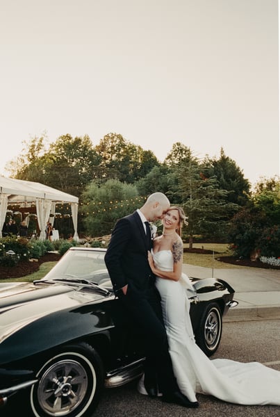 Bride and groom standing close beside the Corvette at sunset, string lights from the reception tent glowing in the background