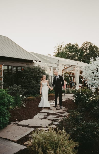 Bride and groom walking hand in hand along a stone garden path at dusk, the lit reception tent behind them