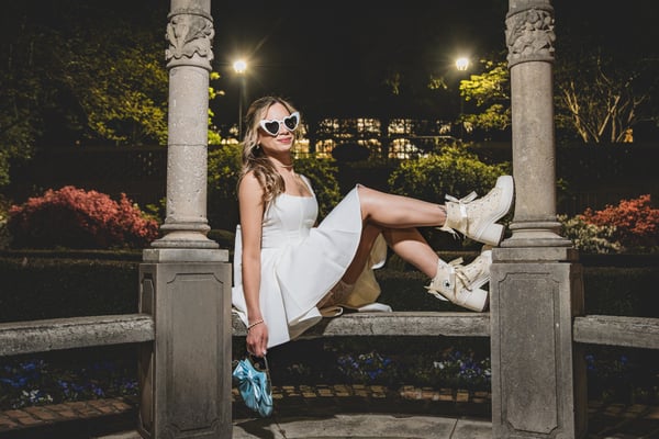 Bride in short white dress seated in the night gazebo, lit azaleas and blue pansies visible in the background, heart sunglasses, blue bag