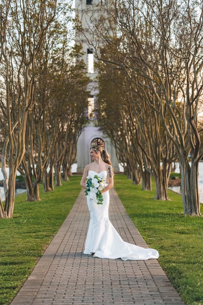 Bride in short white dress seated at night in garden gazebo with azaleas in the background, heart sunglasses and blue bow bag
