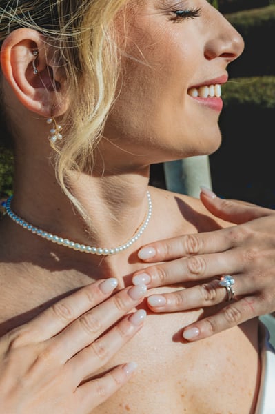 Detail close-up of bride's hands at her collarbone, pearl choker, engagement ring, earring, and laughing upward expression