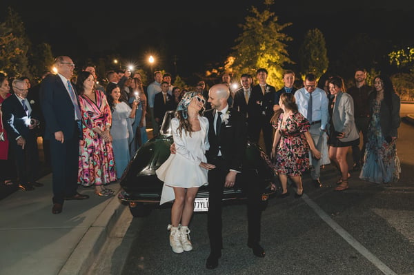 Bride and groom sharing a final laughing moment leaning against the vintage Corvette, surrounded by guests holding sparklers at the send-off, bride in heart sunglasses and blue silk headscarf