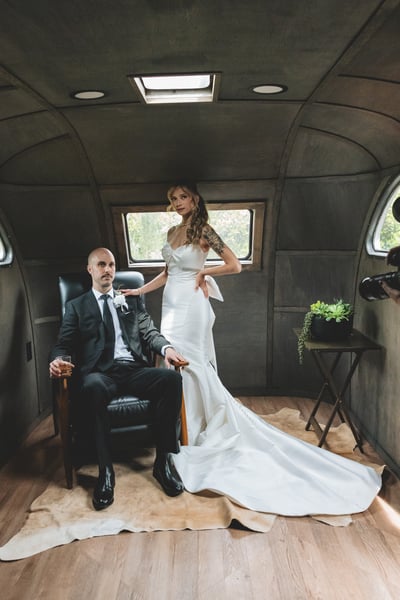 Bride and groom posing together inside a vintage aircraft cabin — groom seated in a leather chair holding a whiskey glass, bride standing beside him in her full gown and train