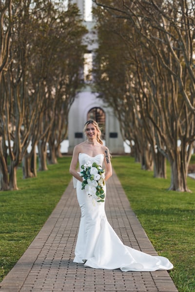 Bride facing the camera on the symmetrical brick tree-lined path, bouquet in hand, smiling in warm afternoon light