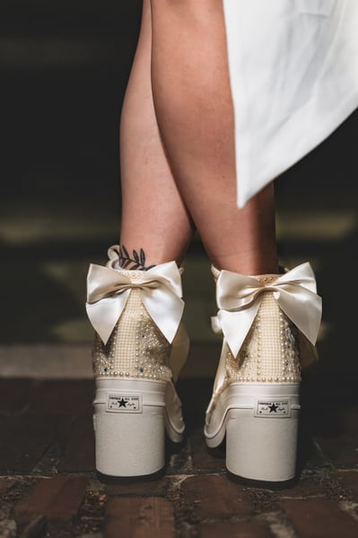 Close-up of backs of both bejeweled platform Converse high-tops with large satin bows, at night, short white dress above