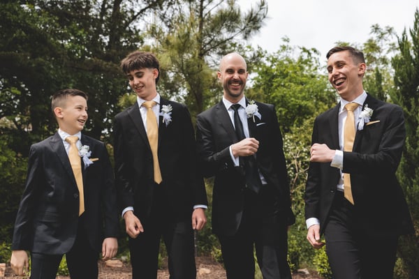 Groom and three groomsmen in dark suits with gold ties walking outdoors and laughing together in a garden setting