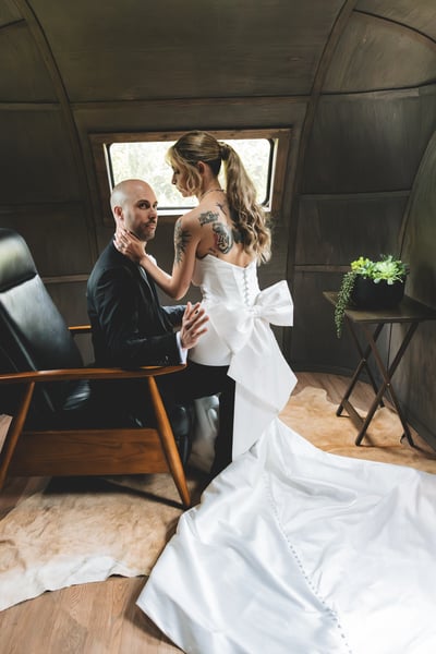 Bride standing over the seated groom inside the vintage aircraft cabin, her hands gently cradling his face as he looks up at her, her bow and tattoos visible from behind