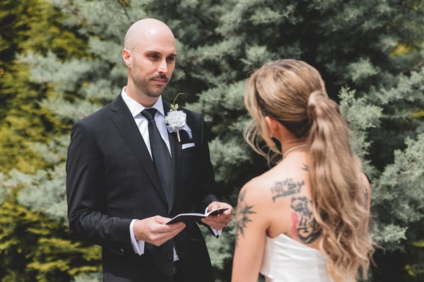 Groom reading his handwritten vows to the bride, who stands facing him with her back to the camera and her gown's large bow visible, set against tall green hedges